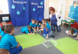 Siedmioro dzieci dotyka rękoma wody w przezroczystym pojemniku. Nauczycielka i pozostałe dzieci obserwuje ich sposoby badania wody.